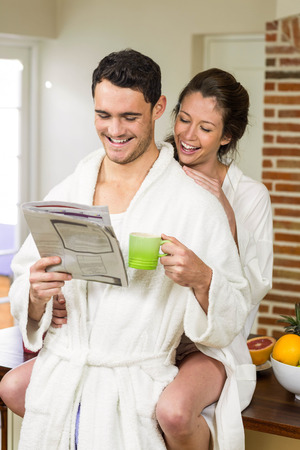 Young couple in bathrobe having tea and reading newspaper in kitchenの写真素材