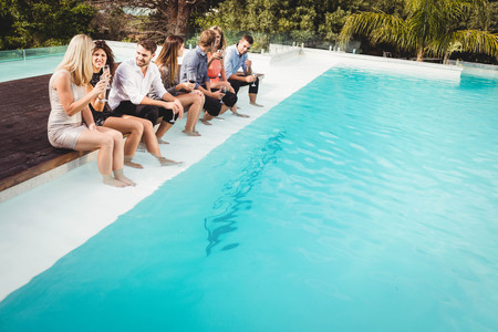 Young people sitting by swimming pool, drinking, having fun, enjoying holiday