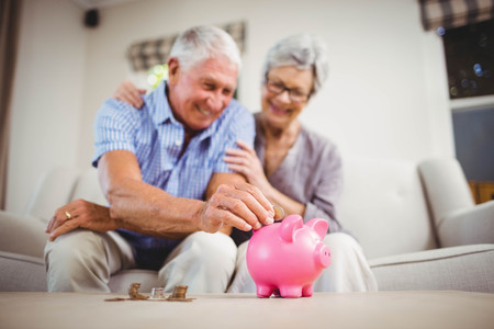 Senior man sitting with woman on sofa and putting coins in piggy bankの写真素材