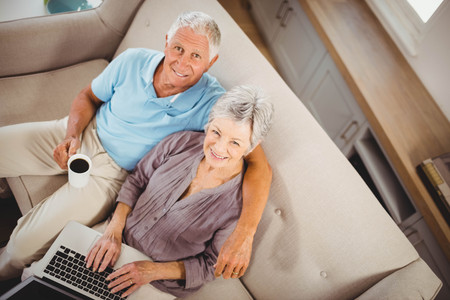 Portrait of senior woman sitting with man on sofa and using laptop in living roomの写真素材
