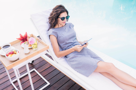 Beautiful young woman relaxing near poolside using digital tabletの写真素材