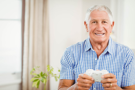 Senior man with joystick looking at camera and smiling in living roomの写真素材
