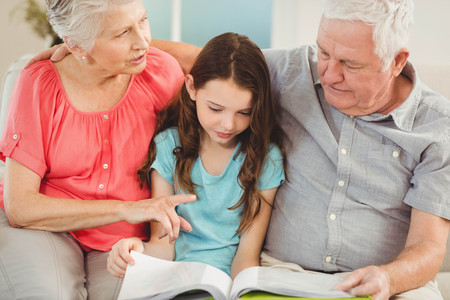 Grandparents and granddaughter sitting on sofa and reading a book with granddaughterの写真素材