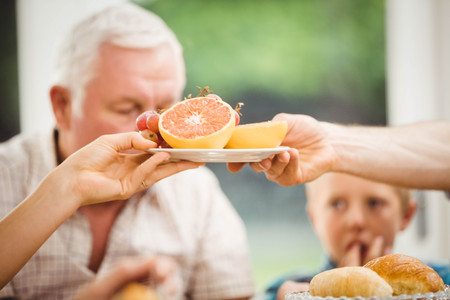 Close-up of hands passing plate of fruits while having breakfast at homeの写真素材