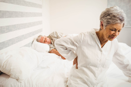 Senior woman suffering from backache sitting on bed in bedroomの写真素材