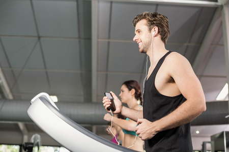 Smiling muscular man on treadmill listening to music at gymの写真素材