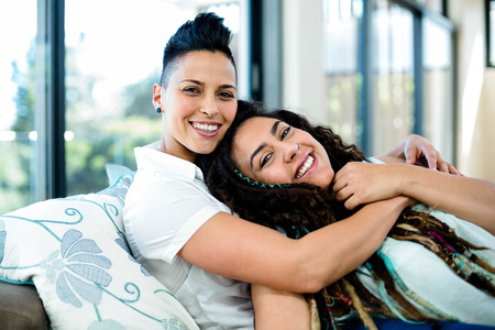 Portrait of smiling lesbian couple embracing and relaxing on sofa in living roomの写真素材