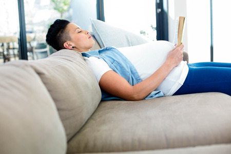 Pregnant woman reading a book while lying on sofa in living roomの写真素材