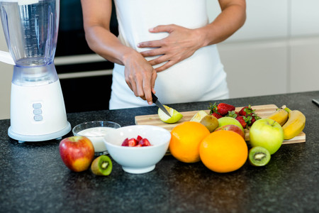 Mid section of pregnant woman touching her belly while cutting fruits on chopping boardの写真素材