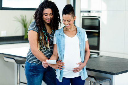 Pregnant lesbian couple looking at sonography report and smiling in kitchenの写真素材