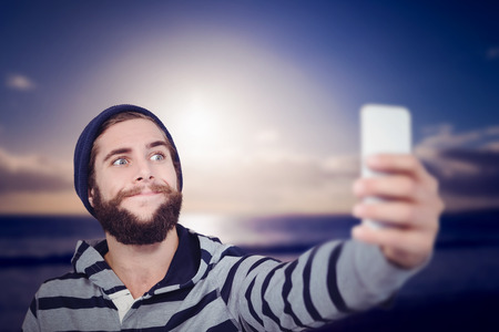 Hipster making face while taking selfie on mobile phone against scenic view of sea against sky