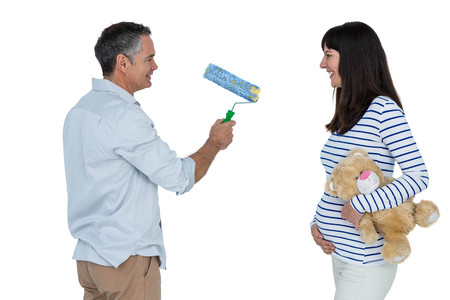 Pregnant woman holding teddy bear while man holding paint roller standing against white backgroundの写真素材