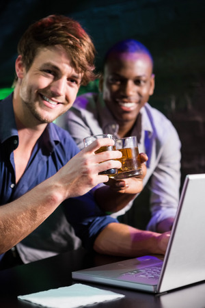 Two men toasting their whiskey glasses at bar counter while using laptopの写真素材