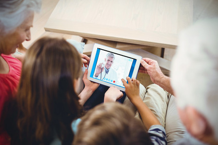 Happy doctor smiling at camera and showing his stethoscope  against family using tabletの写真素材