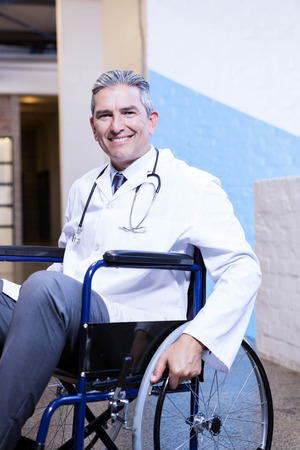 Portrait of happy male doctor sitting on wheel chair in hospitalの写真素材