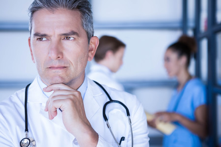 Thoughtful doctor standing with hand on chin in hospitalの写真素材