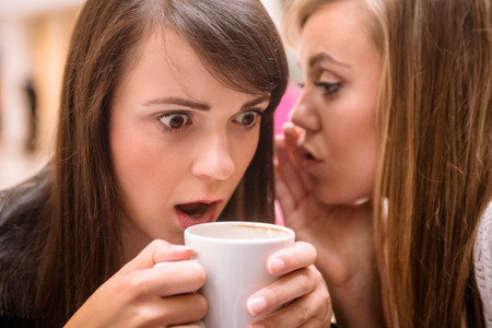 Woman whispering in her friends ears while having coffeeの写真素材