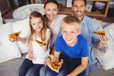 Portrait of smiling family holding pizza slices while sitting on sofa at homeの写真素材