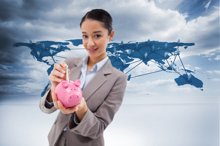 Portrait of a businesswoman putting a bank note in a piggy bank against view of a global mapの写真素材