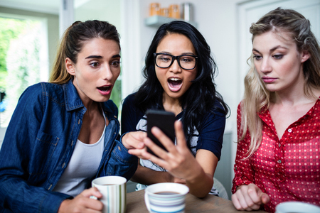 Young female friends shocked while looking in mobile phone at homeの写真素材