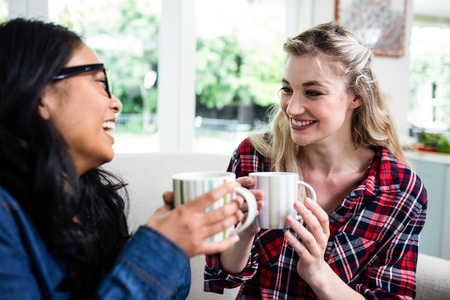 Close-up of cheerful female friends drinking coffee while sitting on sofa at homeの写真素材