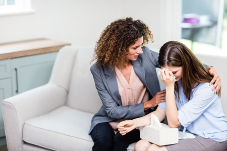 Female psychologist consoling depressed woman while sitting on sofa at homeの写真素材