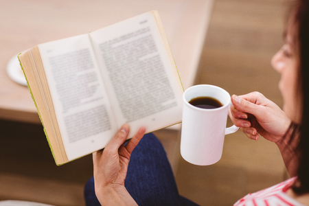 Cropped image of woman reading book while holding coffee cup at homeの写真素材