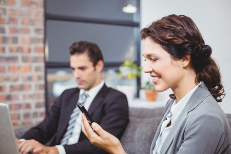 Close-up of businesswoman using mobile phone while male colleague working in backgroundの写真素材