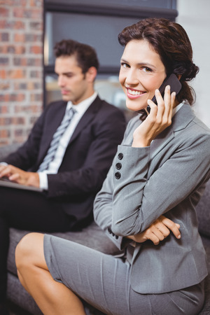 Happy businesswoman using mobile phone while colleague working in backgroundの写真素材