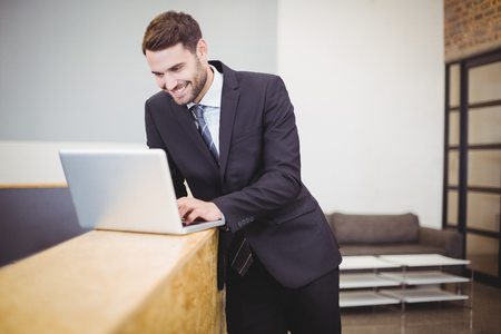 Happy businessman using laptop while leaning on counter at officeの写真素材