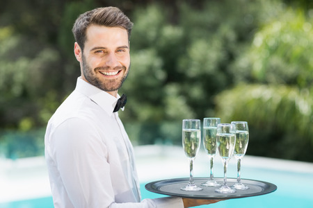 Portrait of happy waiter carrying champagne flutes on tray at poolsideの写真素材