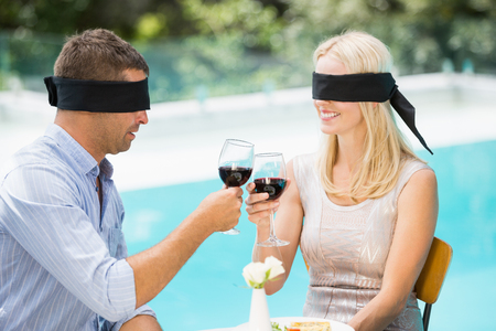 Blindfolded couple toasting red wine while sitting at poolsideの写真素材