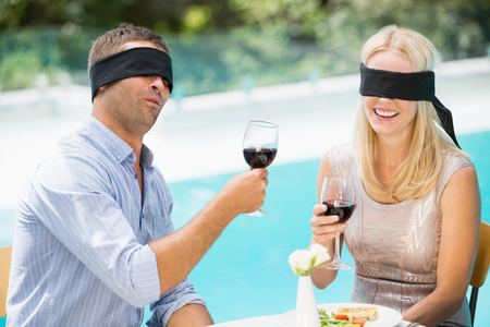 Blindfolded couple holding red wine while sitting at poolsideの写真素材