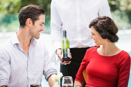 Midsection of waiter showing wine bottle to couple at poolsideの写真素材