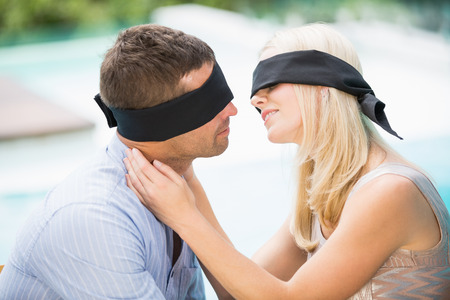 Blindfolded couple kissing while sitting at poolsideの写真素材