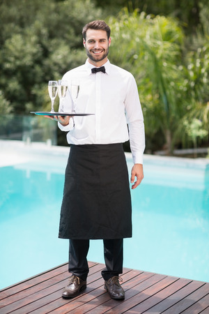 Full length portrait of smiling waiter carrying champagne flutes on tray at poolsideの写真素材