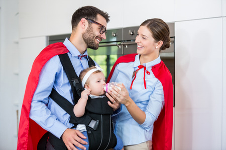 Cheerful couple in superhero costume feeding milk to daughter at homeの写真素材