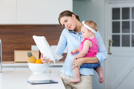 Woman reading documents while carrying baby girl by table at homeの写真素材