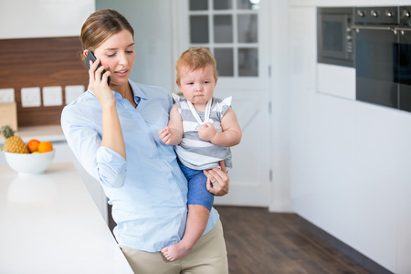 Woman talking on mobile phone while carrying daughter by table at homeの写真素材