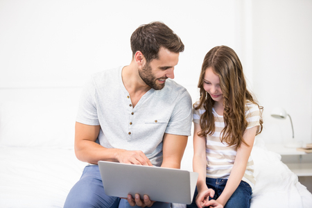 Happy father showing laptop to daughter while sitting on bed at homeの写真素材