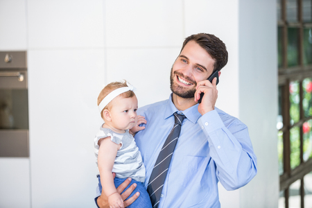 Happy businessman talking on mobile phone while carrying daughter at homeの写真素材