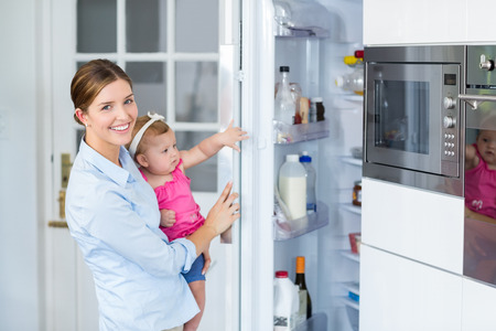 Happy woman opening refrigerator while carrying baby girl at homeの写真素材