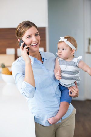 Happy woman using mobile phone while carrying daughter by table at homeの写真素材
