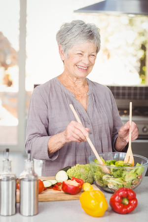 Happy senior woman tossing salad while standing in kitchen at counterの写真素材