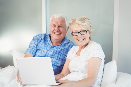 Portrait of happy senior couple with laptop sitting on bed at homeの写真素材