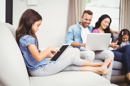 Close-up of girl using digital tablet on sofa while family in background at homeの写真素材