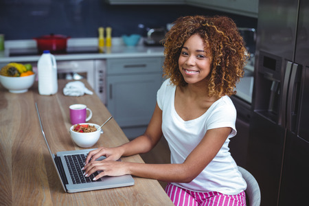 Portrait of happy young woman using laptop at table in kitchenの写真素材