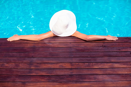 Woman wearing hat leaning on wooden deck by poolside on a sunny dayの写真素材