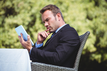 Thoughtful businessman using tablet sitting outside a restaurantの写真素材
