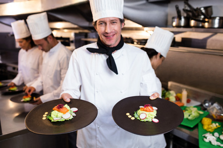Happy head chef presenting his food plates in the commercial kitchen while team working behind himの写真素材